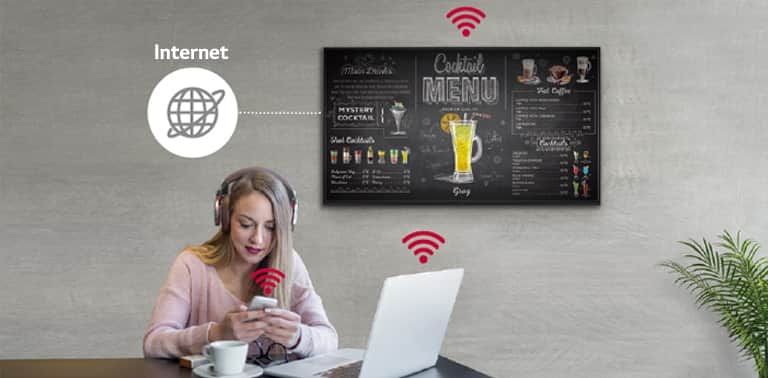 Na ścianie wisi ekran UR3J. Kobieta korzysta z komputera i telefonu komórkowego. Ten obraz pokazuje, że ekran można podłączyć bezprzewodowo do komputera i telefonu komórkowego, który kobieta trzyma w ręku.