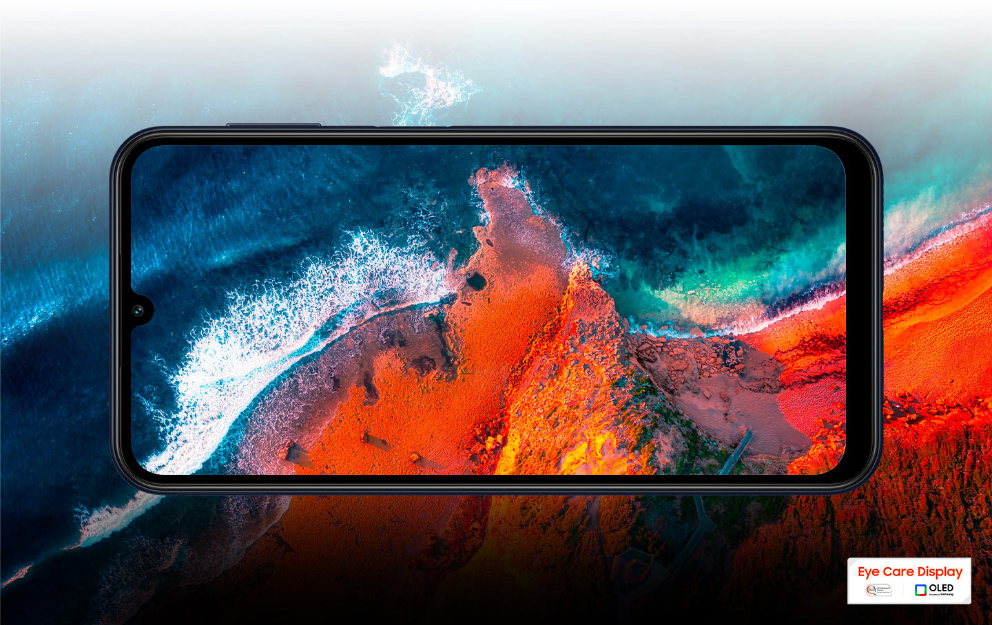 sm-m346-02screen1 Galaxy M34 5G pokazany z lotu ptaka nad plażą i piaskiem. Krajobraz w tle kontynuuje się na wyświetlaczu, przedzielony jedynie ramką urządzenia.