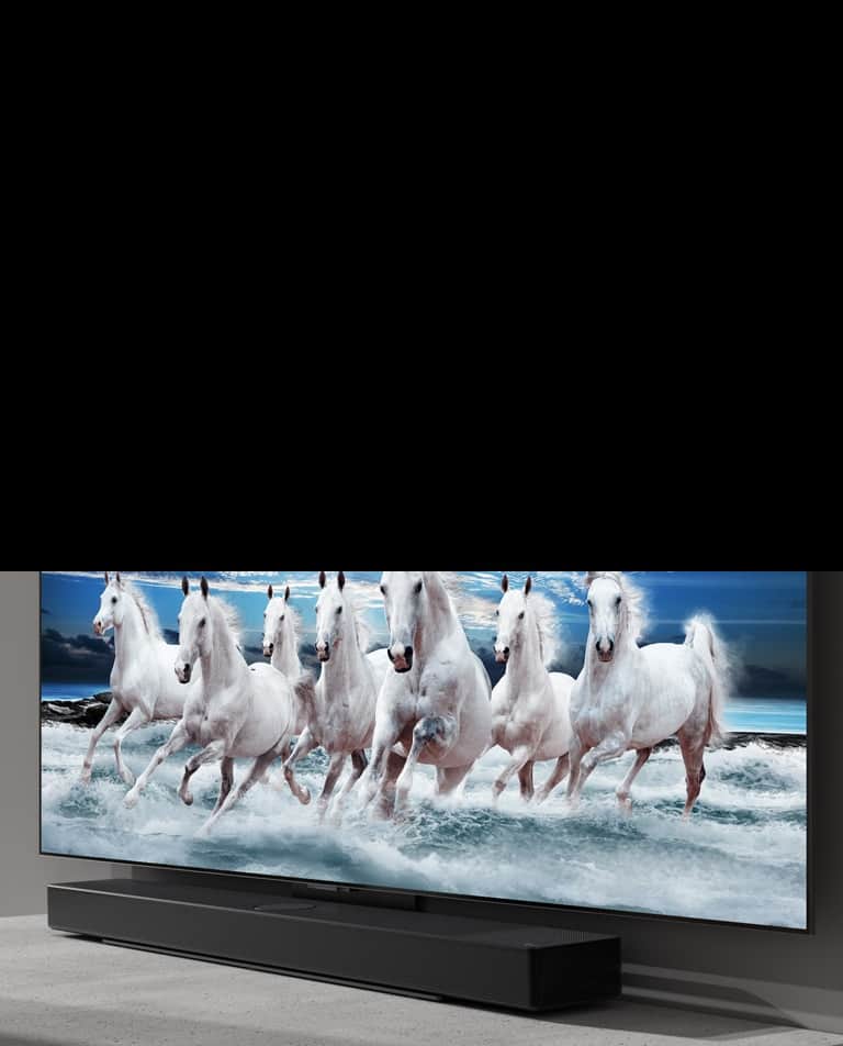 Soundbar i telewizor stoją na białym stole. Na ekranie telewizora widać 7 białych koni.