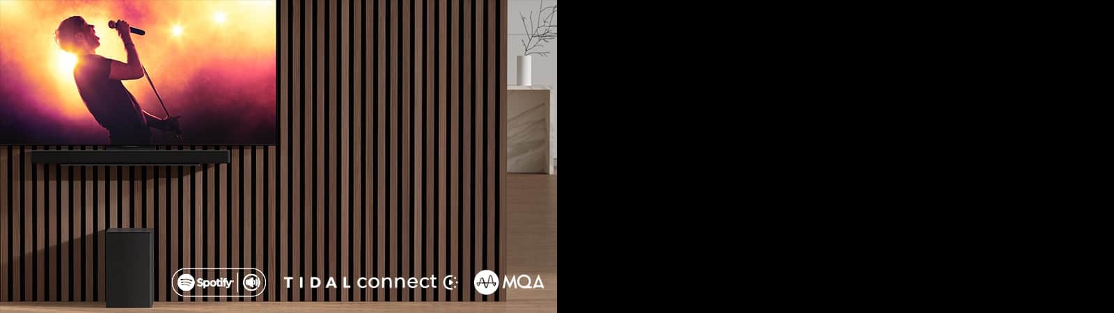Na ścianie wisi telewizor LG OLED serii C, pod nim znajduje się soundbar LG SC9S przymocowany za pomocą specjalnego uchwytu. Pod spodem znajduje się subwoofer. Na ekranie telewizora jest ukazana scena z koncertu.
