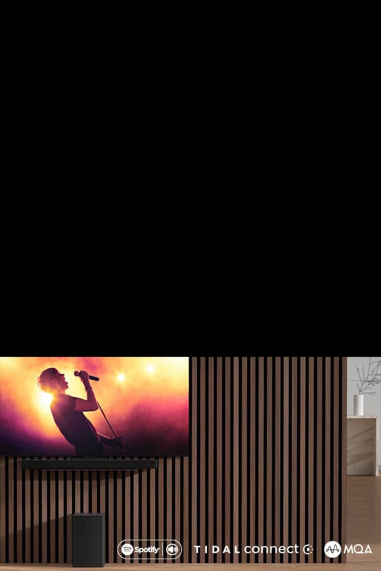 Na ścianie wisi telewizor LG OLED serii C, pod nim znajduje się soundbar LG SC9S przymocowany za pomocą specjalnego uchwytu. Pod spodem znajduje się subwoofer. Na ekranie telewizora jest ukazana scena z koncertu.