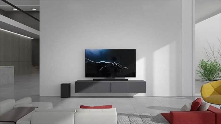 Soundbar stoi na szarej szafce z telewizorem w pokoju dziennym. Czarny bezprzewodowy subwoofer stoi na podłodze po lewej stronie, a z prawej strony obrazu dochodzi światło słoneczne. Biało-czerwona długa kanapa stoi naprzeciwko telewizora i soundbara.