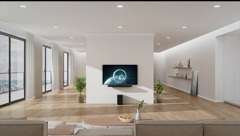 Soundbar i telewizor zawieszone na białej kolumnie na środku pokoju dziennego. Na drewnianej podłodze pod soundbarem stoi czarny woofer. Po lewej stronie znajdują się okna sięgające od podłogi do sufitu ze świetnym widokiem i białymi zasłonami. Po prawej stronie znajdują się drewniane półki z dekoracjami i obrazy na podłodze.