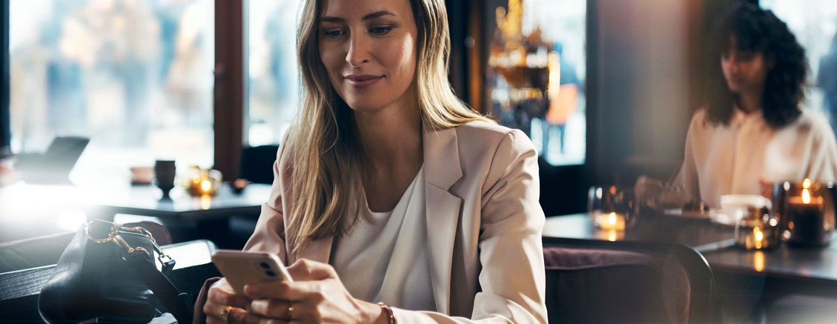 Kobieta w eleganckim stroju siedzi w kawiarni i korzysta ze smartfona przy stoliku.