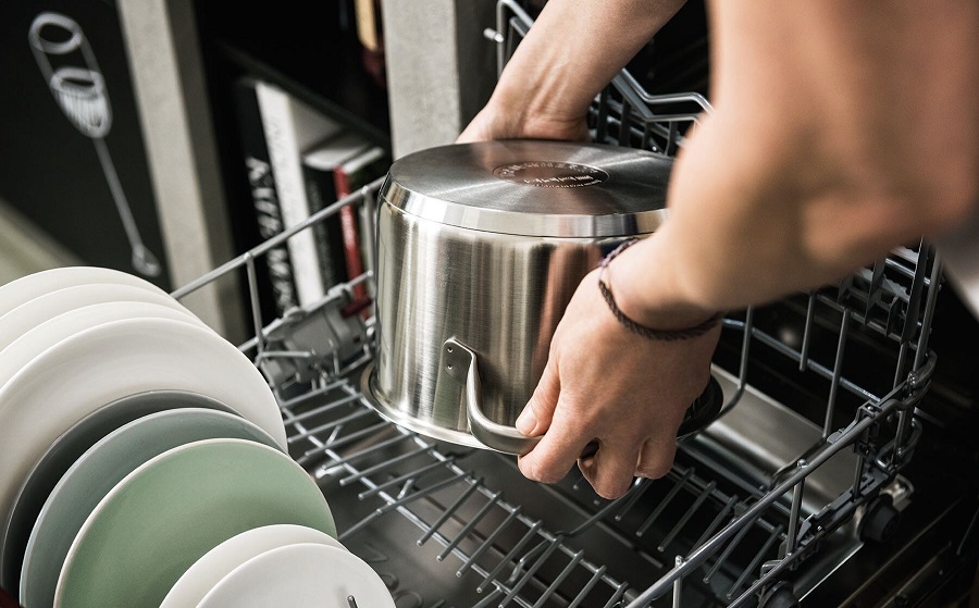 Osoba wkłada duży garnek ze stali nierdzewnej do dolnego kosza zmywarki obok talerzy.
