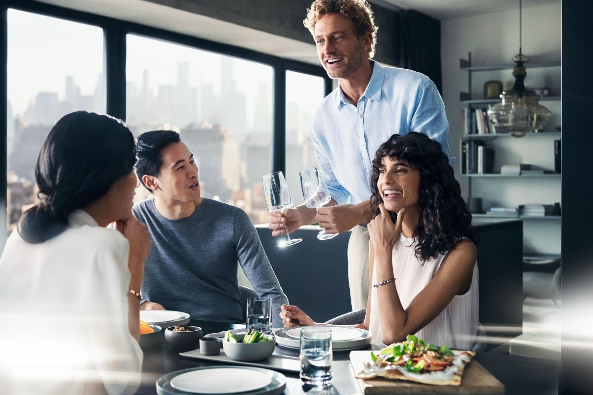 Czwórka znajomych przy stole, uśmiechnięty mężczyzna przynosi kieliszki.